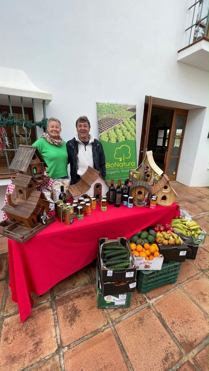 Dos personas sonríen tras una mesa con alimentos, conservas y casitas de madera.